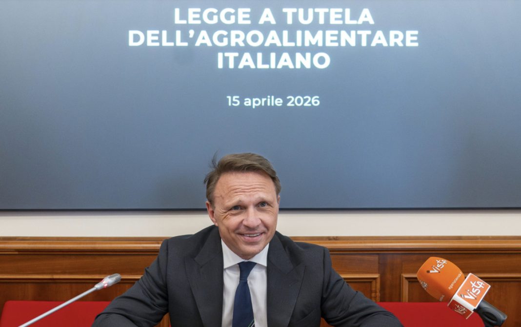 Man in a dark suit at a press conference, with a banner reading 'LEGGE A TUTELA DELL’AGROALIMENTARE ITALIANO' and the date '15 aprile 2026' behind him.