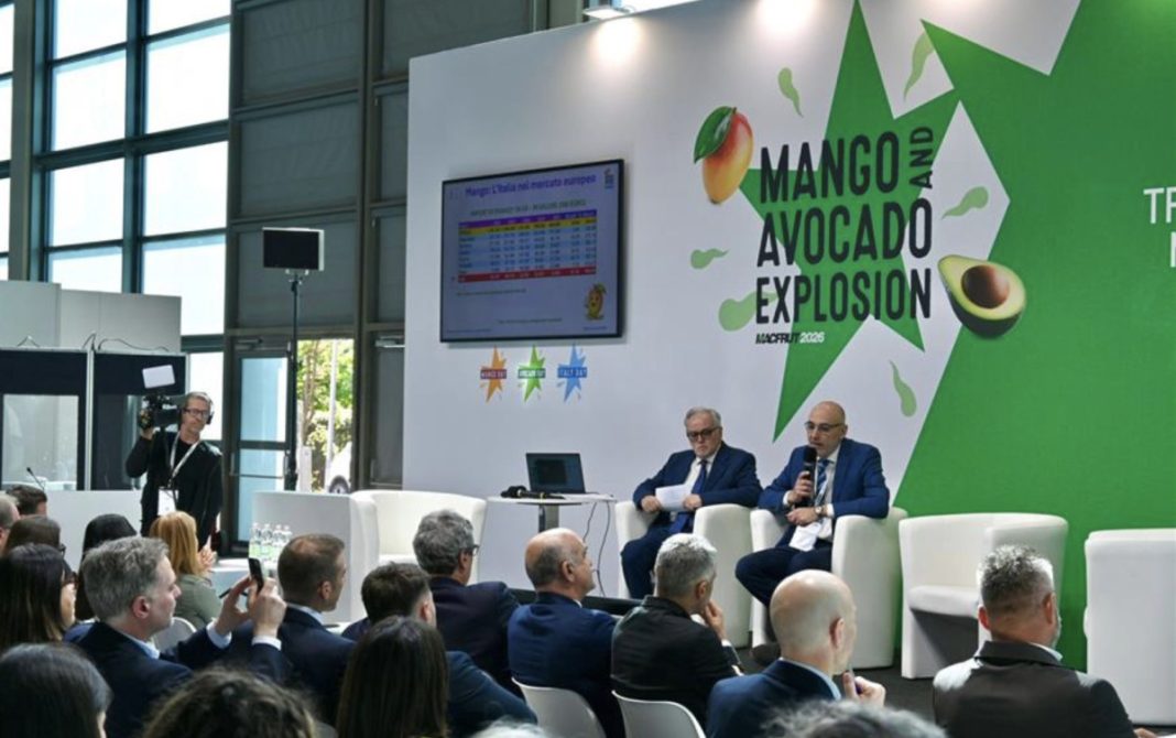 Panel discussion at a conference with two men in suits on stage, mango avocado explosion banner behind them on a bright green wall.