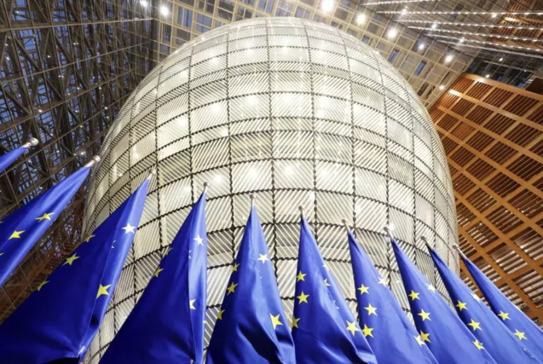 Blue European Union flags with yellow stars fluttering in front of a large glass dome atrium in a modern building.