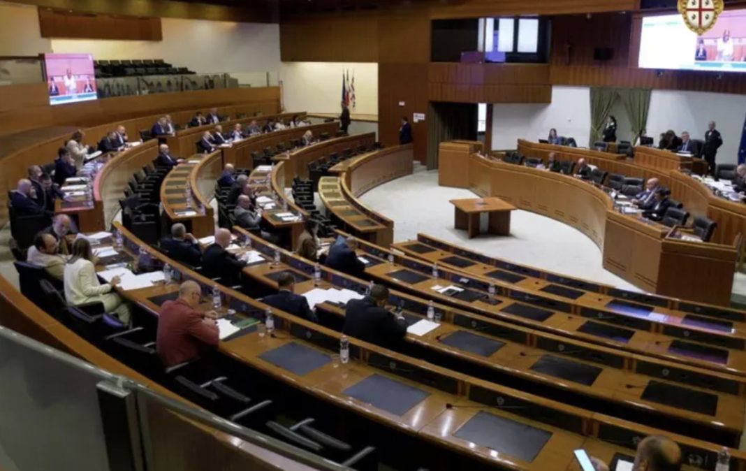Legislative chamber with curved wooden desks and politicians seated at their benches during a session, papers and bottles on desks, flag visible in the background.