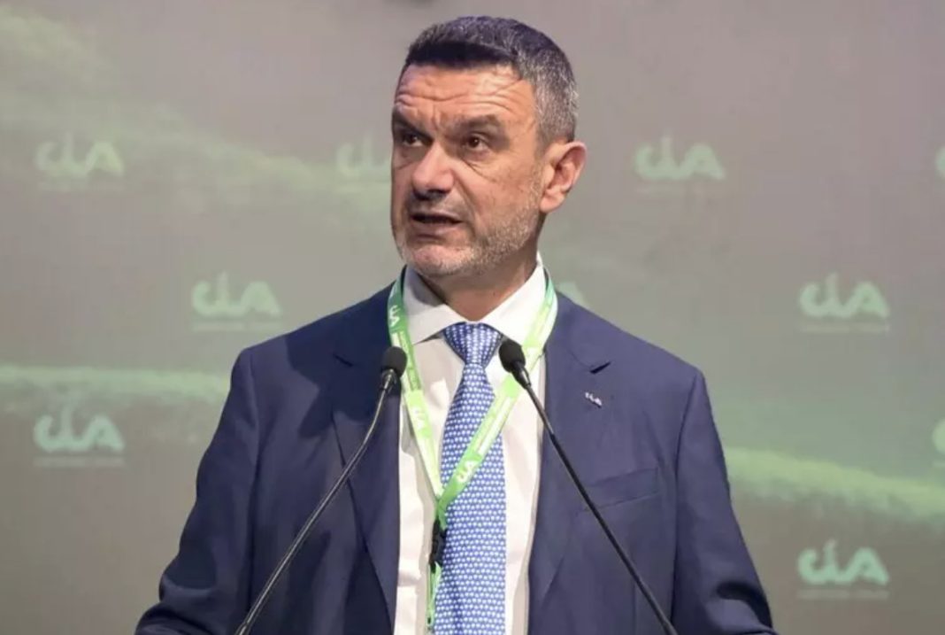 Man in a navy suit speaks at a podium with two microphones, wearing a green lanyard at a conference.