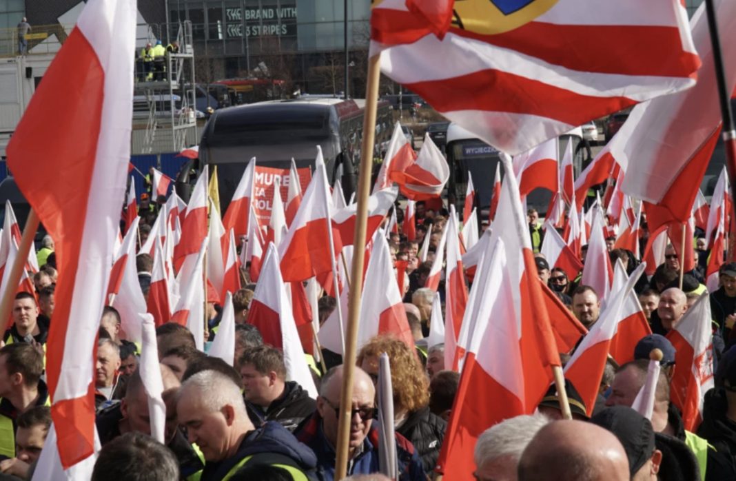 Large crowd of protesters waving red-and-white Polish flags in an urban street.