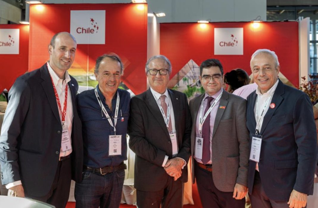 Five men in business attire posing together at a Chile booth with red backdrop and lanyards at a conference venue.