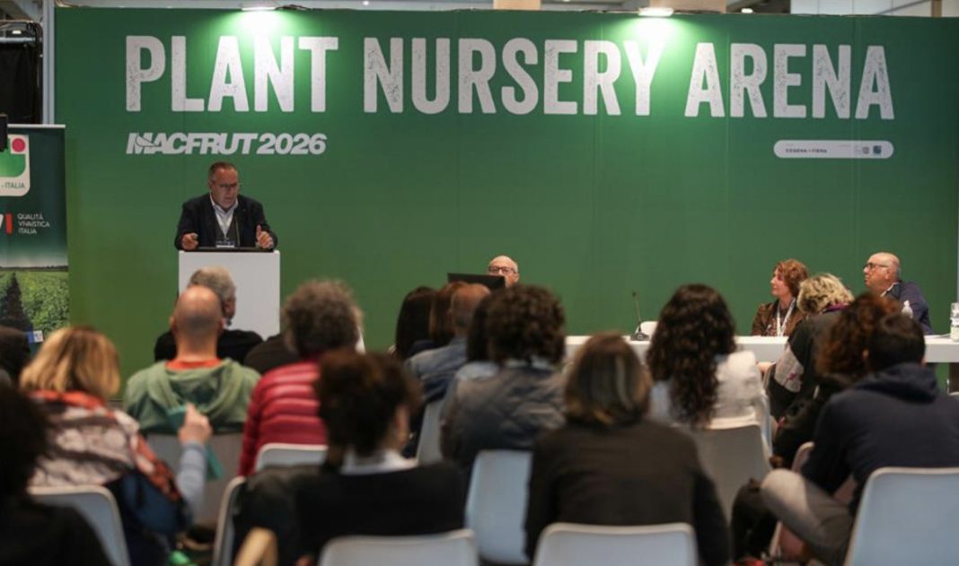 Man in a suit speaks at a podium on a stage labeled 'PLANT NURSERY ARENA' with a seated panel and audience in front of a green backdrop.