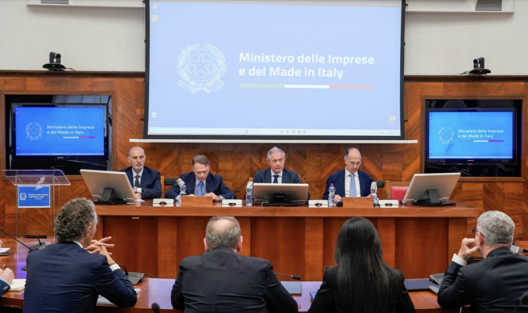 Panel of five officials seated at a wooden dais in a conference room, with a large screen behind reading 'Ministero delle Imprese e del Made in Italy' and two side monitors showing the same title. Audience in foreground.