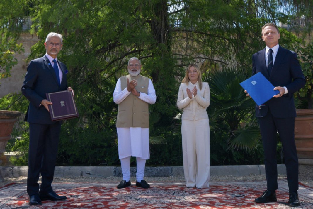 Four dignitaries stand on a red outdoor carpet; two men in suits hold folders while two others clap nearby.