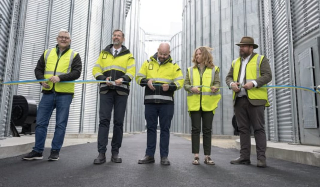 Five people in high-visibility vests cut a blue ribbon at an industrial facility opening, flanked by metal silos and a paved area.