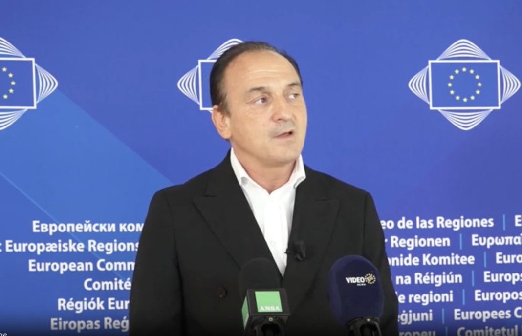 Man in a black suit giving a press conference in front of a blue EU backdrop with microphones in front of him