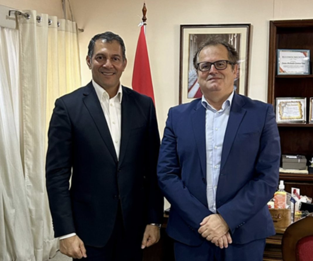 Two men in business suits pose for a photo in a formal office, with a red flag and framed artwork behind them.
