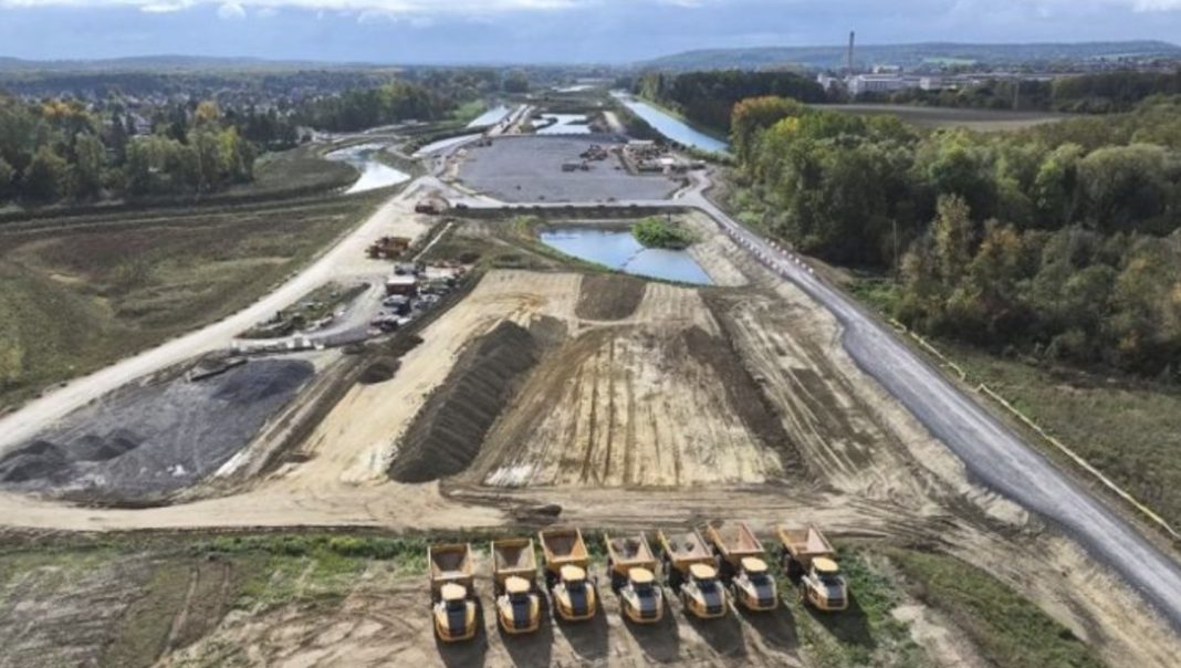 Aerial view of a construction site with dirt piles, graded earth, and a row of orange dump trucks at the front, with roads and water features in the background