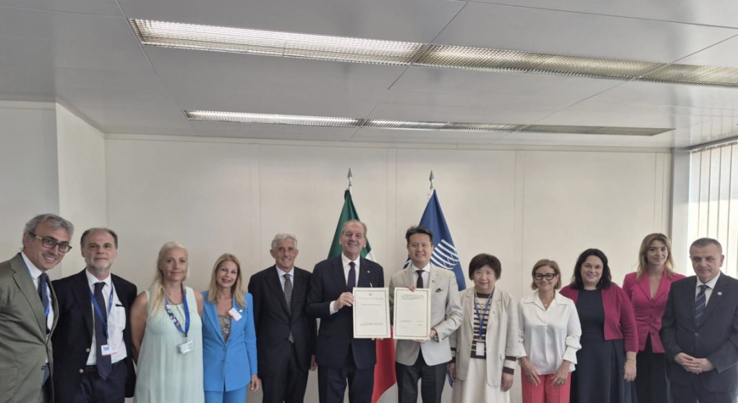 Group of twelve professionals in formal attire posing for a signing ceremony; two men in the center hold up signed documents, flanked by colleagues, with two flags (green and blue) in the background behind them.