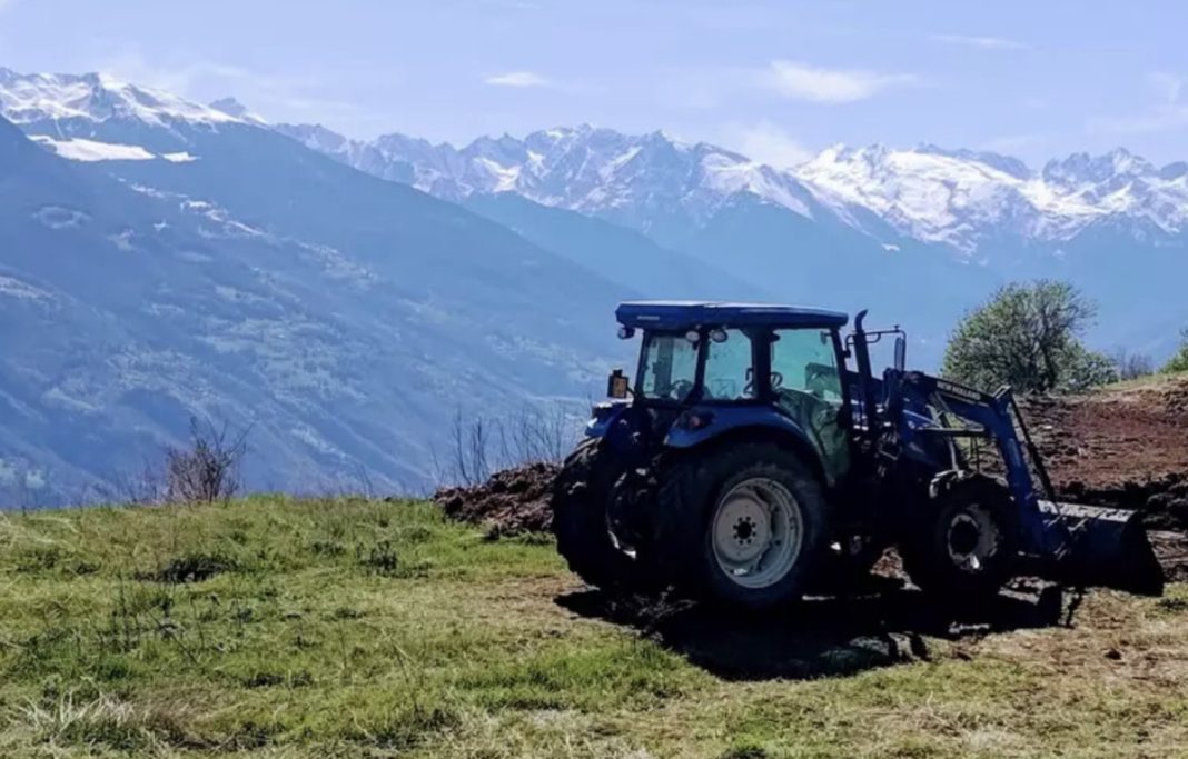 Blue tractor with a front loader parked on a grassy hillside, with snow-capped mountains in the distance inside a blue sky.