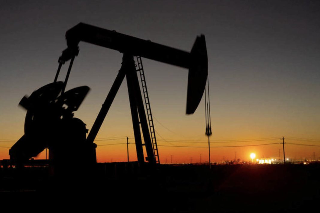 Silhouette of an oil pumpjack at sunset with an orange horizon and distant lights behind it.